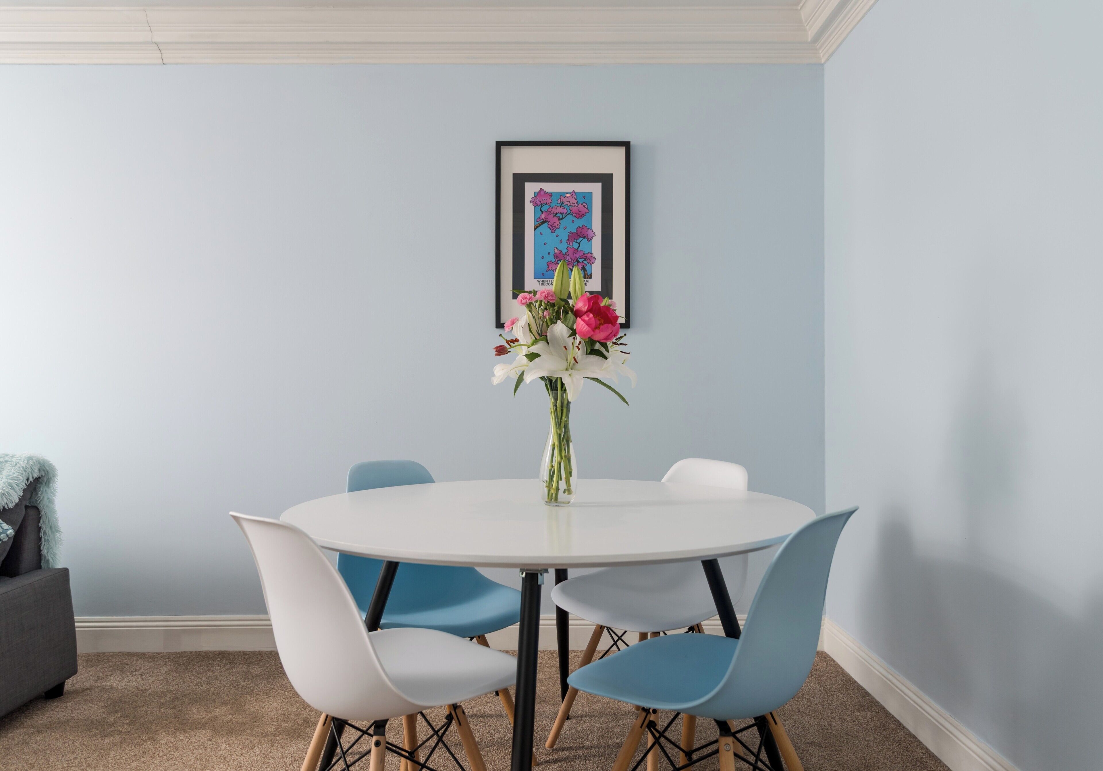 A room with a round white table, blue chairs, a vase with flowers, and a framed picture on the wall.