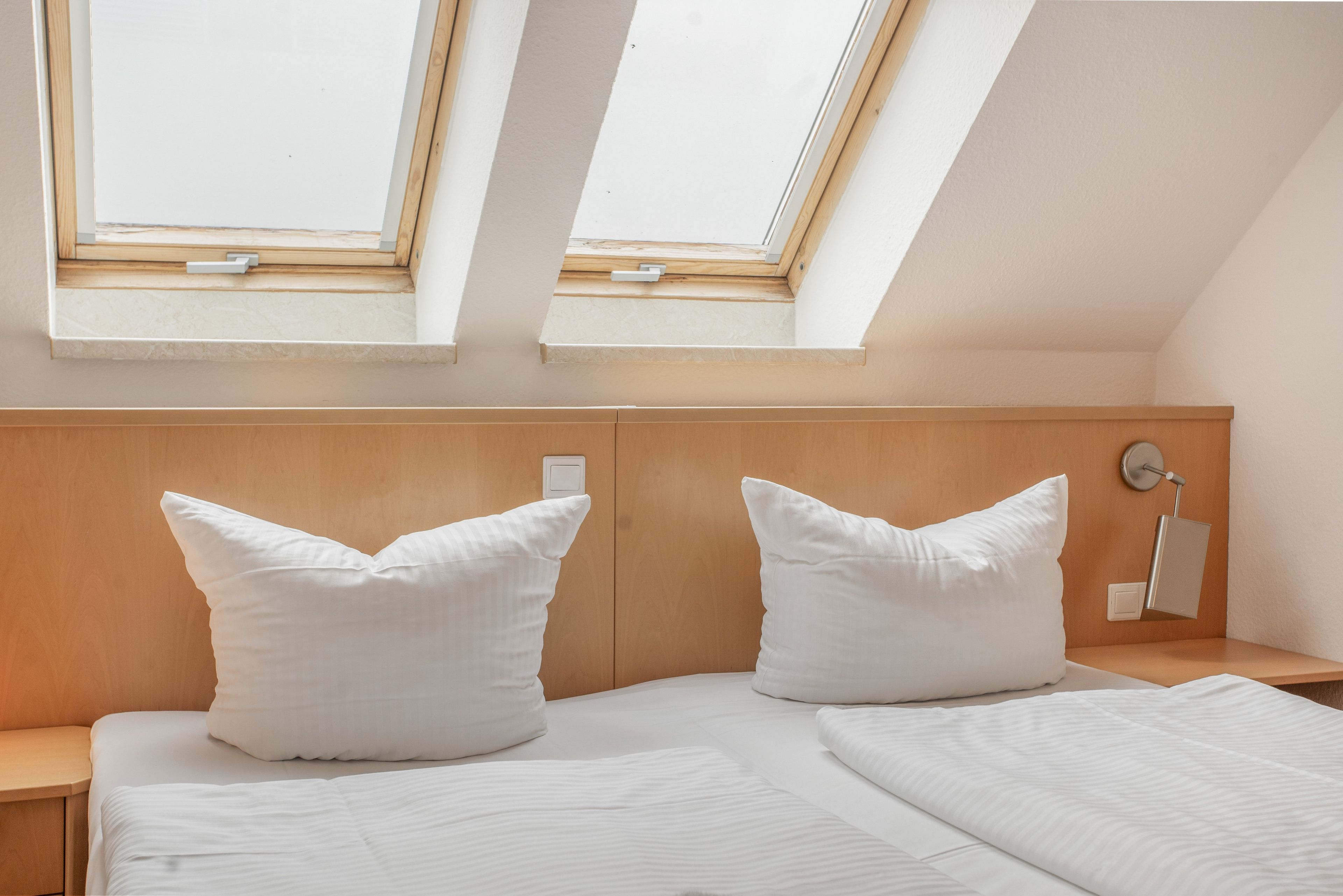 Ein Schlafzimmer mit schrägen Wänden, zwei Dachfenstern, einem Holz-Kopfteil, einem Nachttisch, einer Lampe und zwei Kissen.