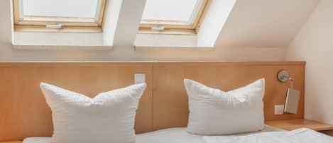 Un dormitorio con techo inclinado, dos tragaluces, cabecero de madera, mesita de noche, lámpara y dos almohadas.
