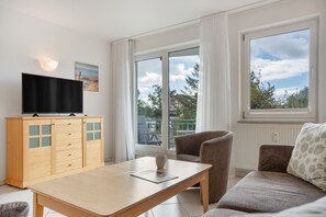 A modern living room with a flat-screen TV on a wooden cabinet, a sofa, a coffee table, and large windows offering a view of the outdoors. - Apartment 'Ava' with Balcony and Wi-Fi (Zempin)