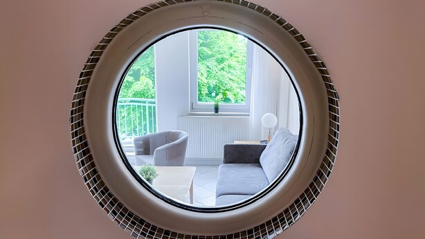 Une pièce dotée d’une lucarne circulaire, d’un canapé, d’un fauteuil, d’une petite table et d’une fenêtre donnant sur un paysage verdoyant.