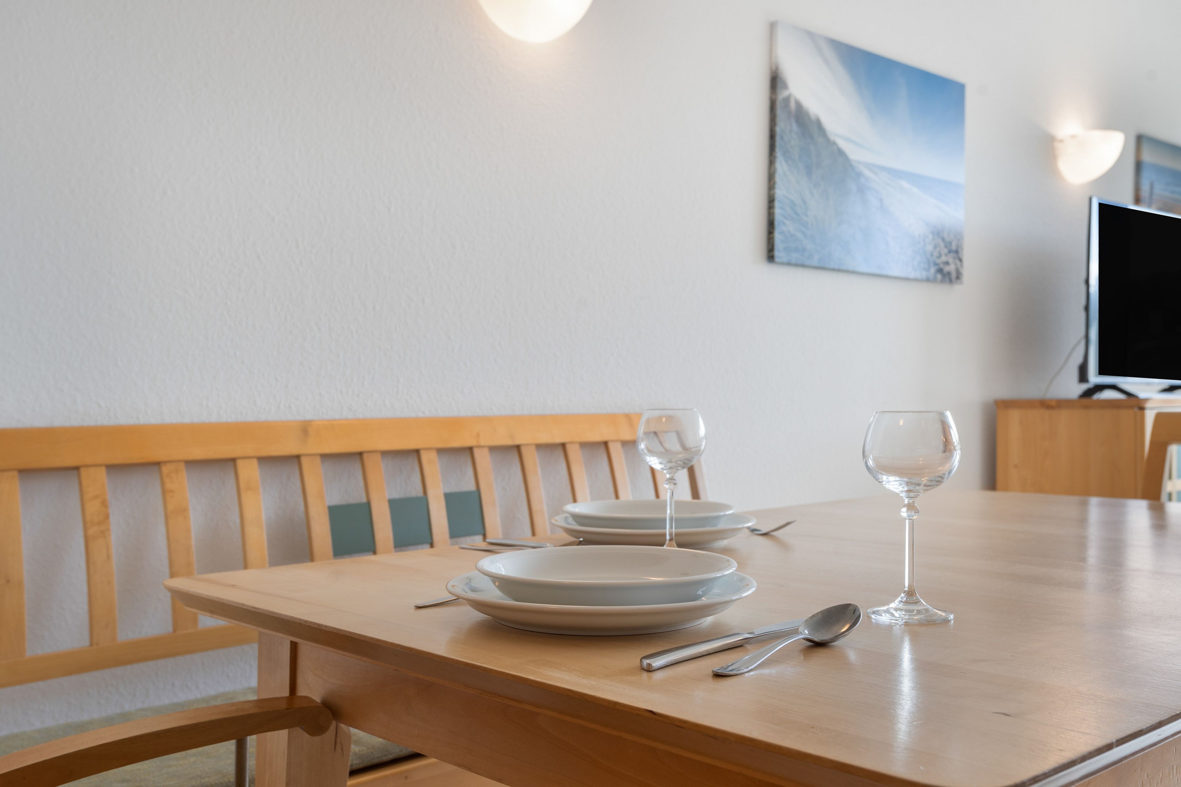 Een eettafel met borden, bestek en glazen, een houten bank en een televisie op een meubel.