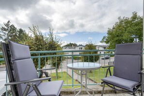 Ein Balkon mit zwei Stühlen und einem kleinen Tisch, der auf eine Straße mit Häusern und Bäumen blickt.
