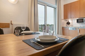 A dining area with a table set for two, a view of the outdoors through a window, and a kitchen area with a microwave. - Apartment "Tine" with Balcony and Beach Chair in Summer (Zempin)