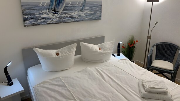 En snyggt bäddad säng med vita sängkläder, en sänggavel och kuddar. På väggen hänger en tavla med segelbåtar, och i rummet finns en golvlampa och en stol med en kudde.