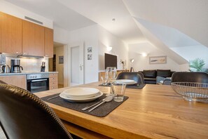 A modern kitchen with wooden cabinets, a dining table set with plates, glasses, and cutlery, and a living area with a sofa and a coffee table. - Apartment "Tine" with Balcony and Beach Chair in Summer (Zempin)