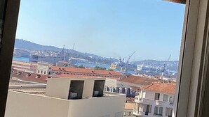 Blick aus einem Fenster auf ein Gebäude mit blauem Himmel, einen Hafen mit Booten und sanfte Hügel in der Ferne.