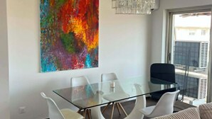 Ein modernes Wohnzimmer mit einem Glastisch, weißen Stühlen, einem beigen Sofa und einem farbenfrohen, abstrakten Gemälde an der Wand.