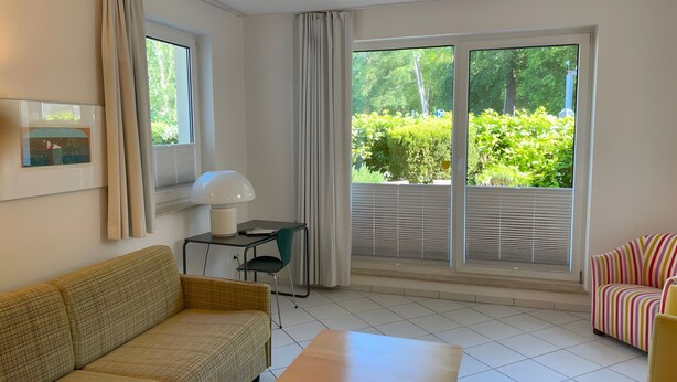 Ein Zimmer mit einer Couch, einem kleinen Tisch mit Lampe, einem Sessel und einem großen Fenster mit Jalousien.
