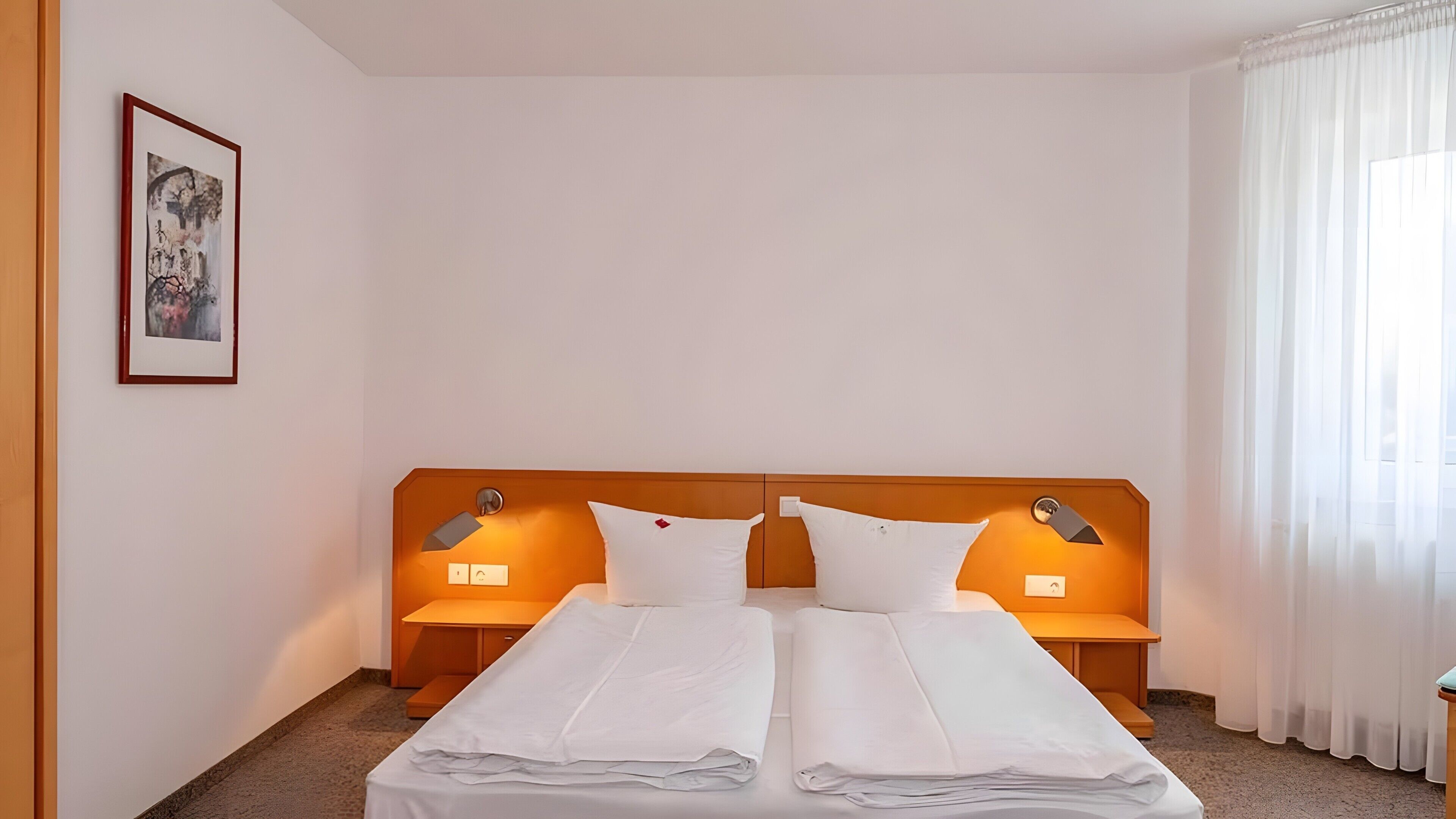Une chambre d’hôtel avec un lit double, deux tables de chevet, une tête de lit, un tableau encadré au mur et une fenêtre avec des rideaux.