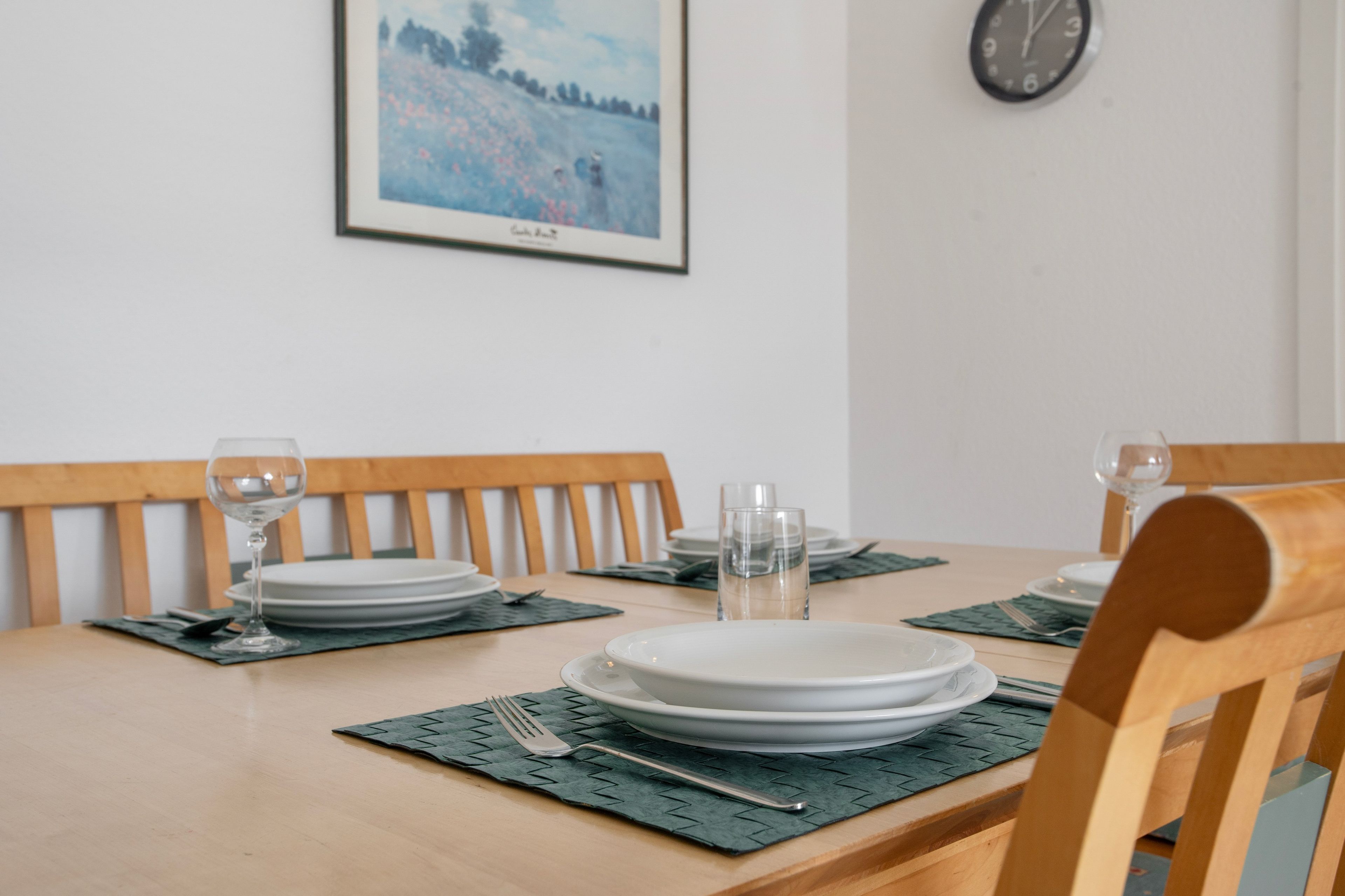 A dining table set with plates, glasses, and cutlery, with a framed picture and a clock on the wall.