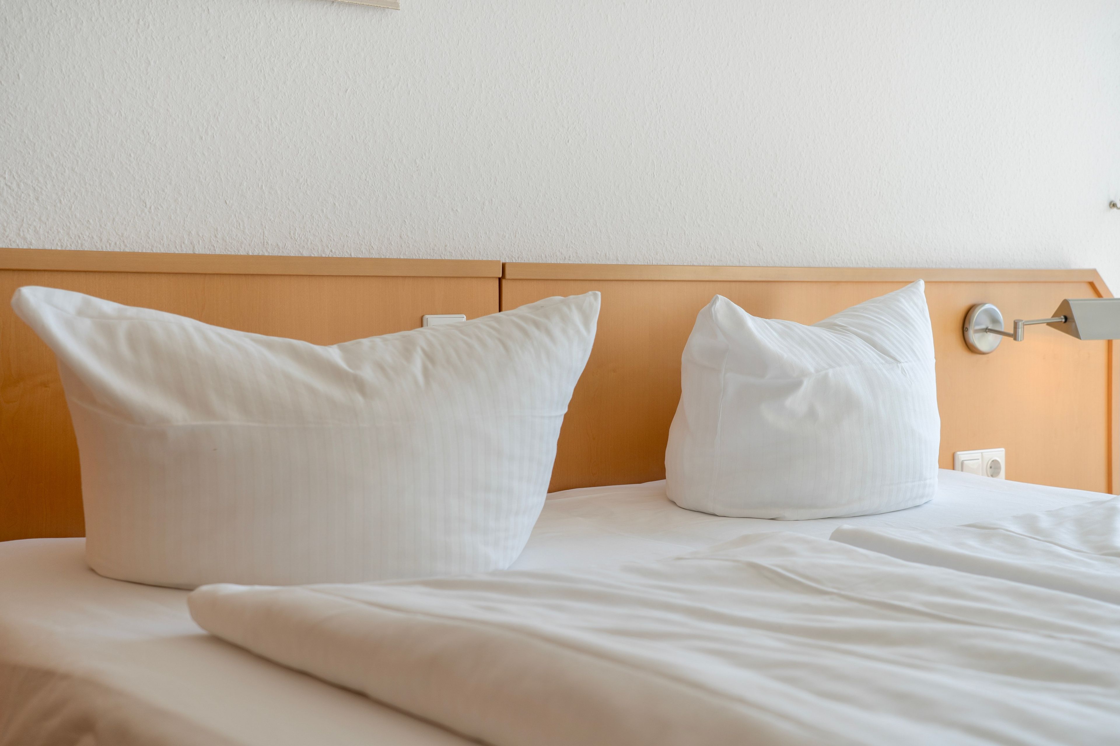 Una cama bien hecha con ropa blanca y almohadas, cabecera de madera y una lámpara fijada a la pared.