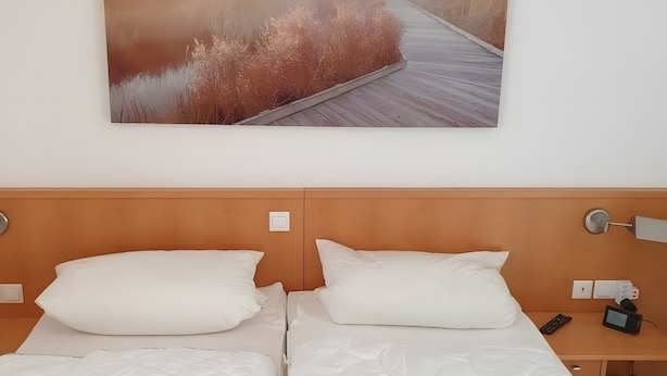 A hotel room with a wooden headboard, two beds with white linens, a bedside table with a phone and a digital device, and a wall-mounted painting of a natural scene. - Apartment 'Sonnenblick' with Balcony and Wi-Fi (Zempin)