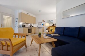 Ein gemütlicher, kompakter Wohnbereich mit einer Couch, einem Holztisch, einem gelben Sessel, einer Küchenzeile mit Geräte und einem kleinen Esstisch.