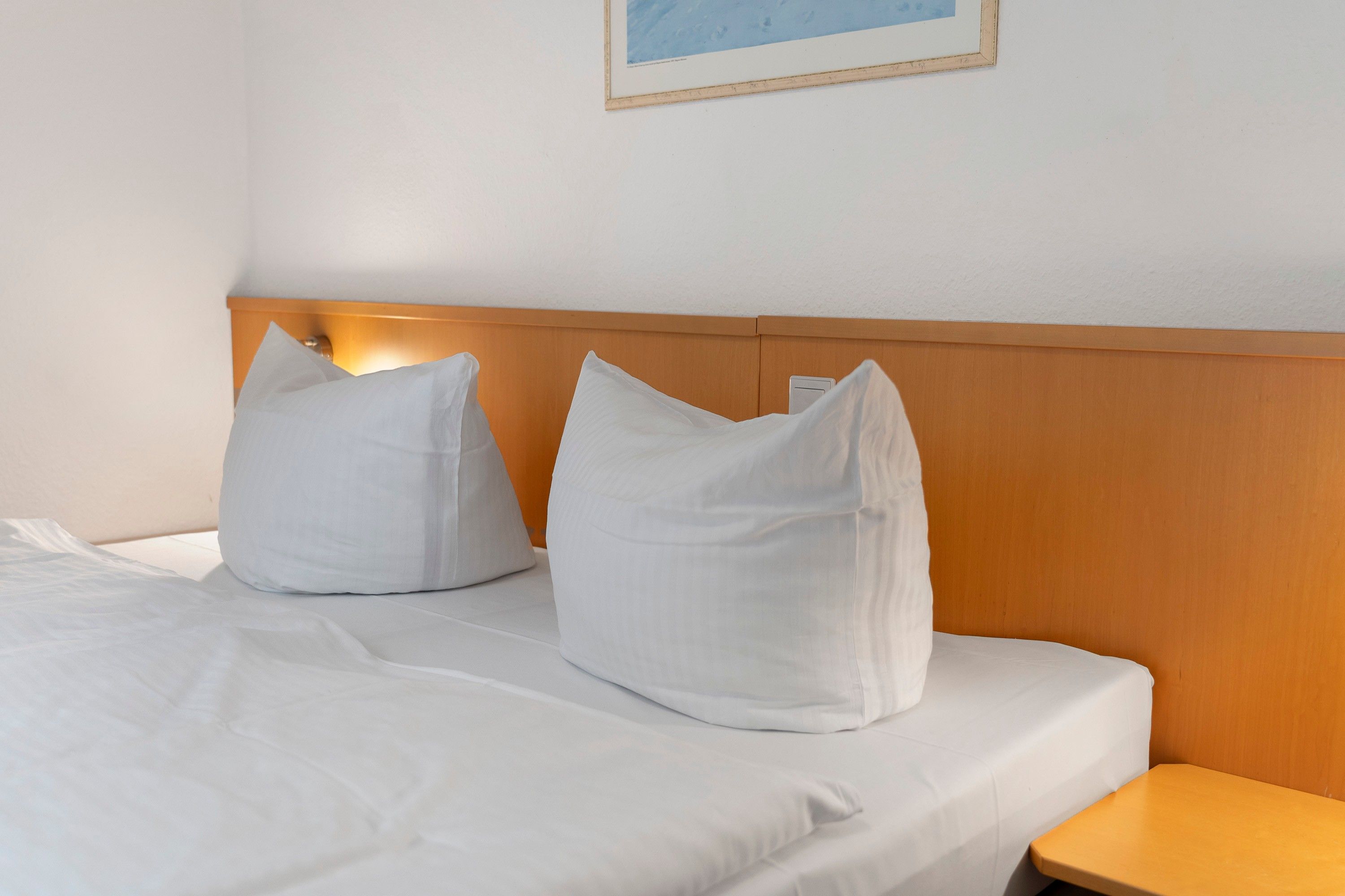 A hotel room with a bed, pillows, a nightstand, and a framed picture on the wall.
