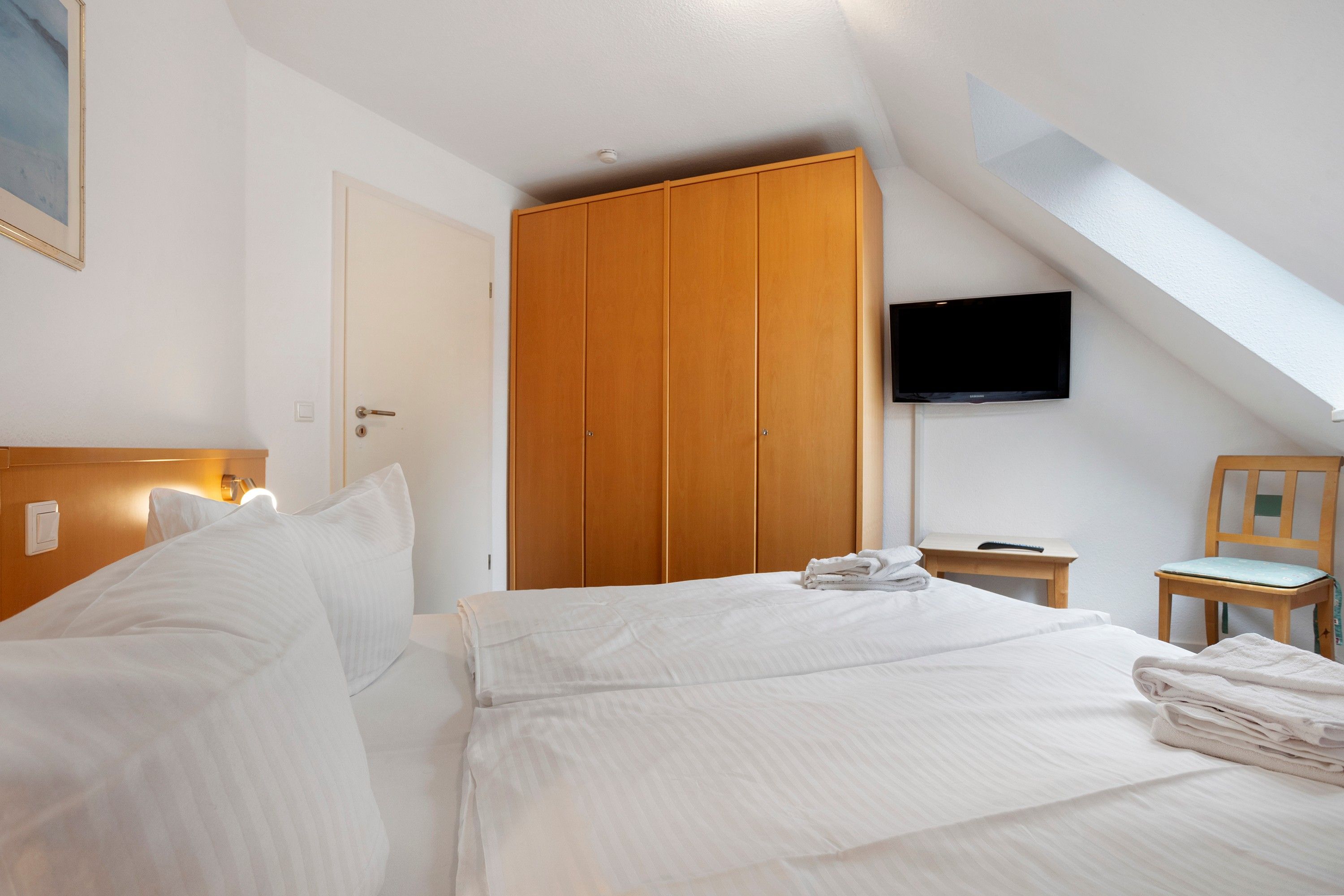 A hotel room with a bed, a wooden wardrobe, a television mounted on the wall, and a small table with a chair.