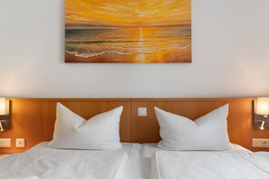 Habitación de hotel con cama, dos almohadas, cabecero, lámparas de noche y un cuadro de una puesta de sol sobre el océano.