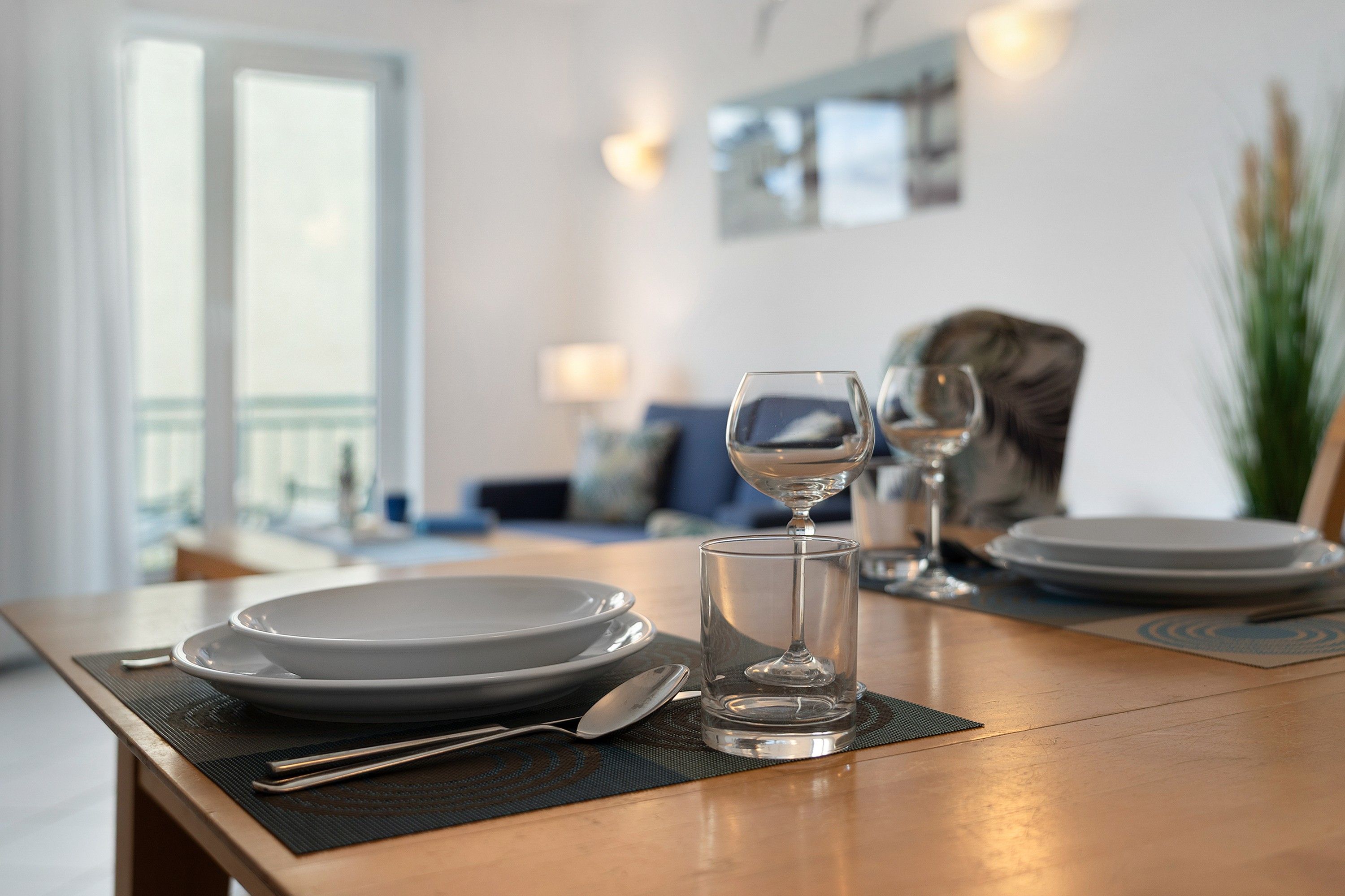 Una mesa de comedor dispuesta con platos, cubiertos y vasos, con una sala de estar al fondo.