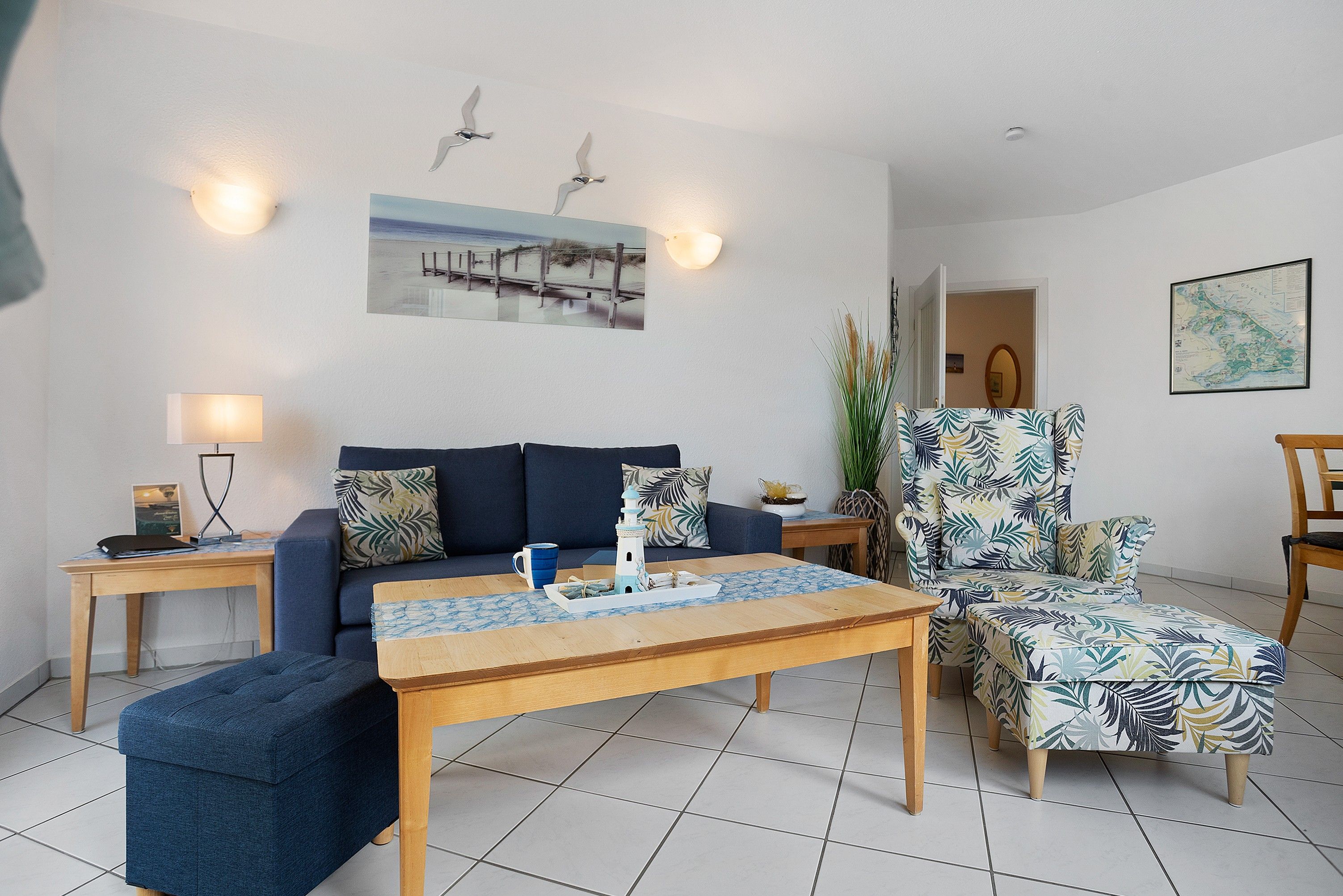 Un salon comprenant un canapé, un fauteuil, une table basse et une lampe. Il y a un tableau au mur et une carte accrochée au mur.
