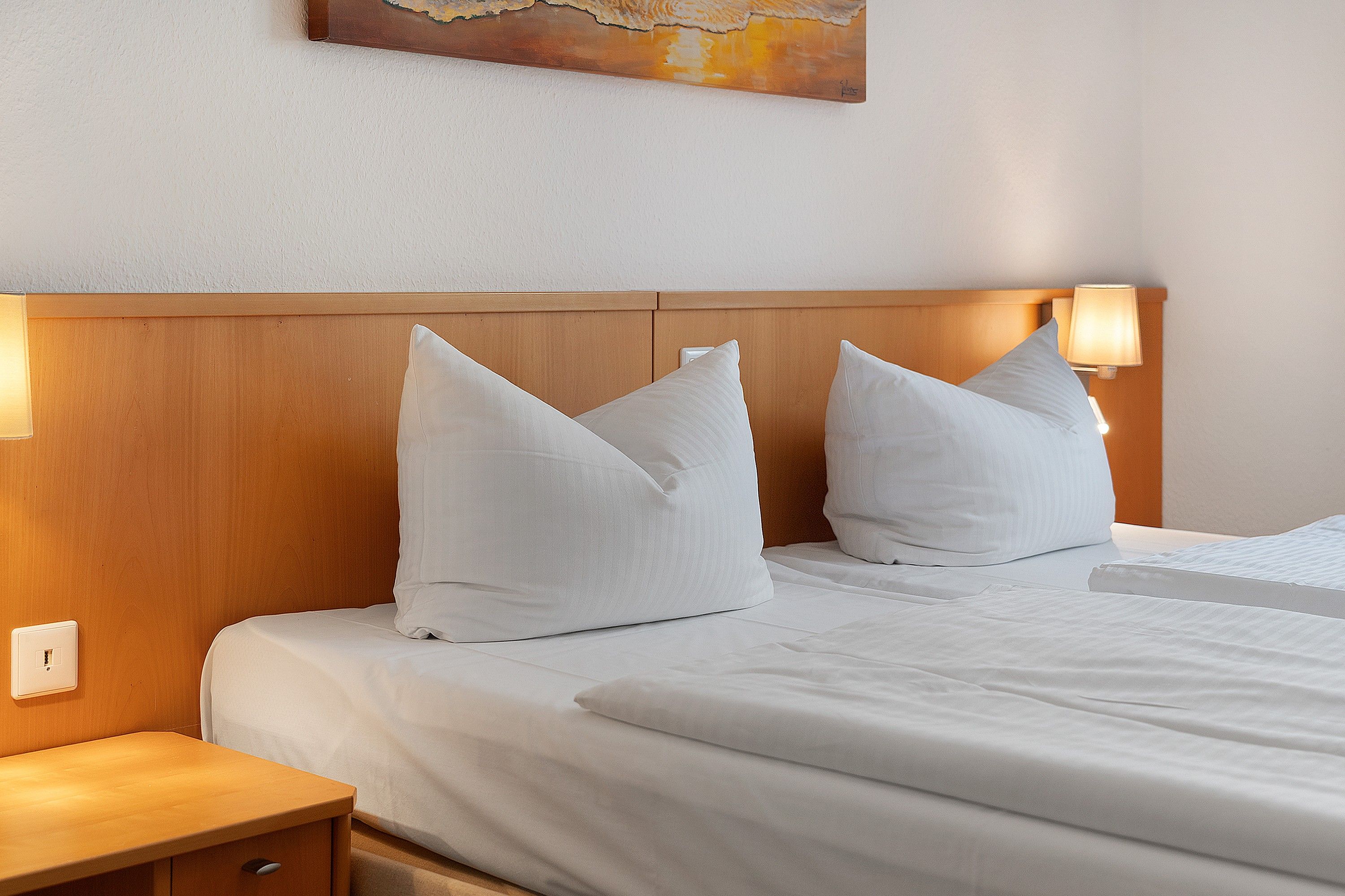 A hotel room with a wooden headboard, two bedside lamps, a bed with white bedding, and a painting on the wall.