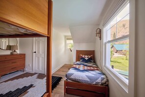 A bedroom with a bunk bed, a wooden dresser, a bedside table, a lamp, a patterned rug, and a window with a view of a house and landscape.