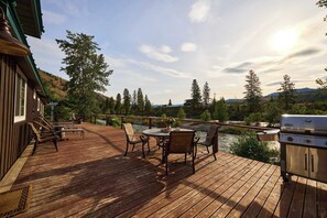 A wooden deck with outdoor furniture, a grill, and a view of a river and forest.