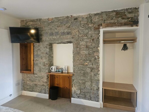 A room with a stone wall, a mounted television, a wooden shelf with a kettle, and a coat rack.