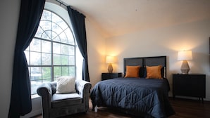 A bedroom with a large window, a bed with dark bedding, two bedside lamps, a chair, and a nightstand.