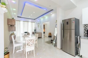 A modern kitchen with a stainless steel refrigerator, a dining area with a white table and chairs, and a living area with a sofa and a coffee table. - Indigo | Designer 2BR/2BA Apartment, Gated Complex, Pool & Beach Access - Juan Dolio (Playa Juan Dolio)