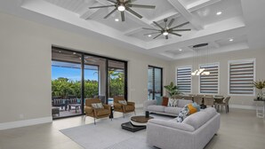 Un amplio salón con un sofá grande, sillones y una mesa de comedor. Hay ventiladores de techo, ventanas grandes y vistas al exterior.
