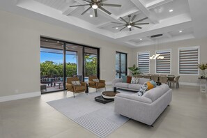 Un vaste espace de vie comprenant un grand canapé, des fauteuils et une table à manger. On y trouve également des ventilateurs de plafond, de grandes fenêtres et une vue sur l’extérieur.