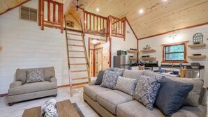 Interior acolhedor de cabana com sala de estar, sofá, mesa de centro, cozinha e um quarto no mezanino com escada de madeira.