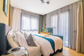 A hotel room with a large bed, a bedside table with a vase and a tray of fruit, and a view of the city through sheer curtains. - DAR DYAFA VIP (Agadir)