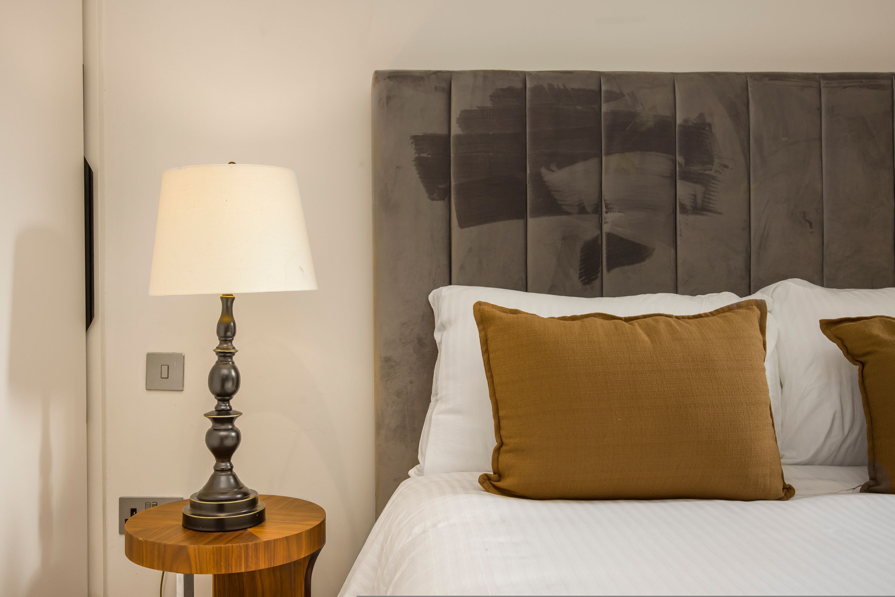 Een net opgemaakt bed met witte en bruine kussens, een rond houten nachtkastje met een lamp, en een wand met een structuurpaneel.