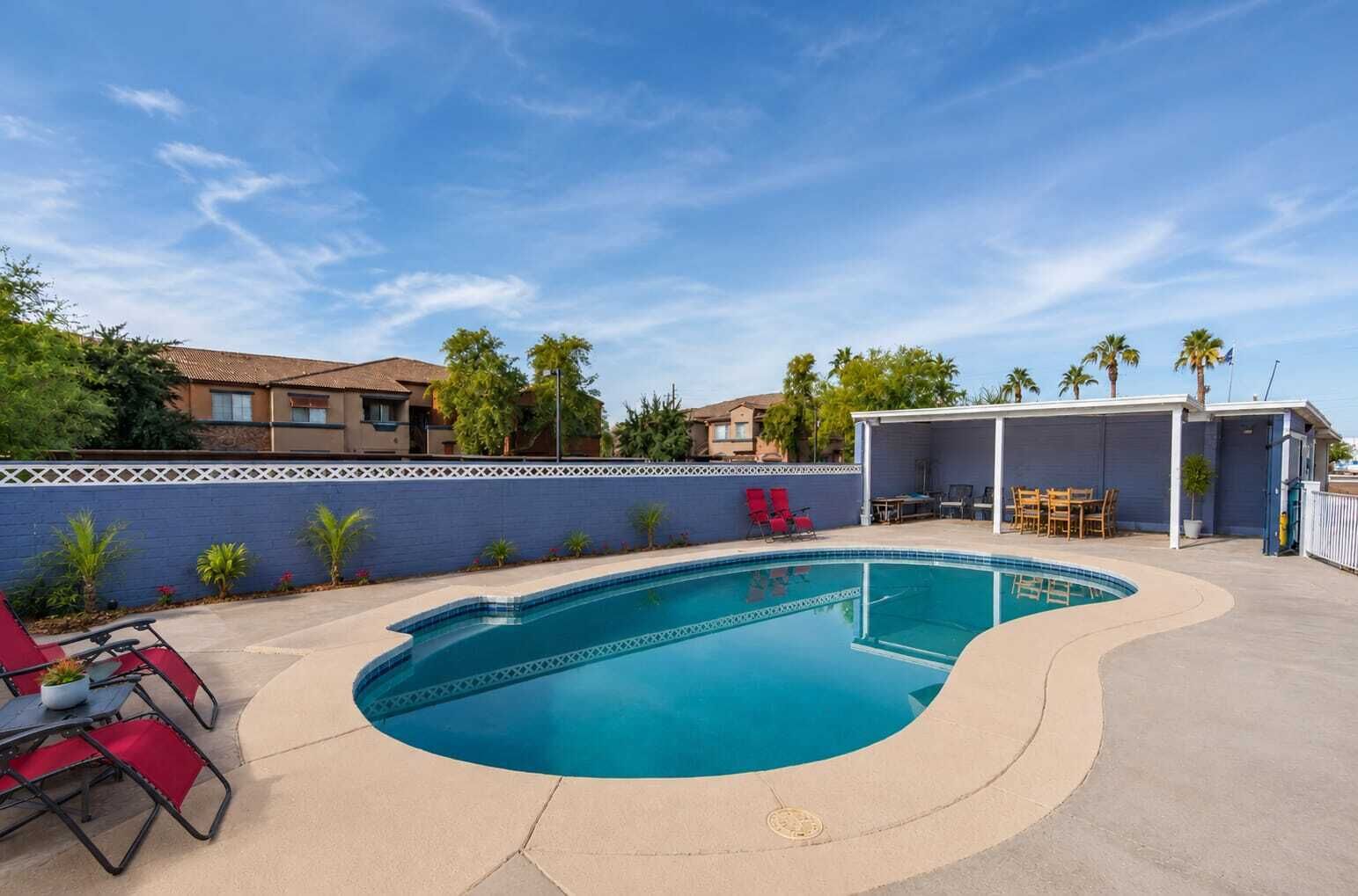 Ein Pool mit gewölbter Kante, umgeben von einer blauen Wand und einer überdachten Terrasse mit Gartenmöbeln.