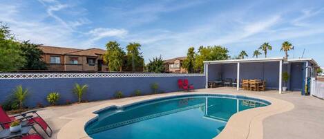 Ein Pool mit gewölbter Kante, umgeben von einer blauen Wand und einer überdachten Terrasse mit Gartenmöbeln.