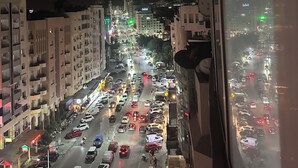 Ein nächtliches Stadtbild mit Gebäuden, einer belebten Straße und Blick durch ein Fenster.