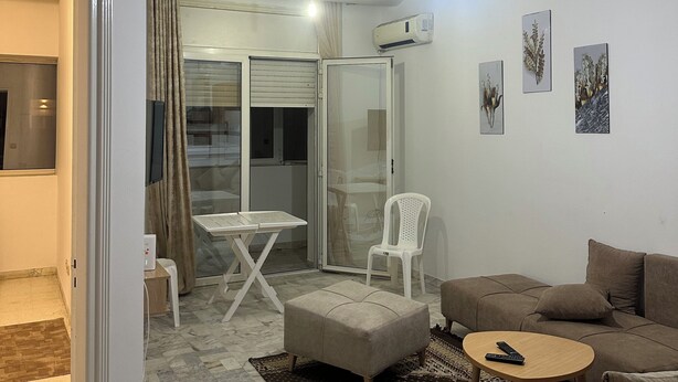 Ein modernes Wohnzimmer mit einer Couch, einem Hocker und einem kleinen Tisch. Eine Glastür führt zu einem Balkon mit Tisch und Stühlen.