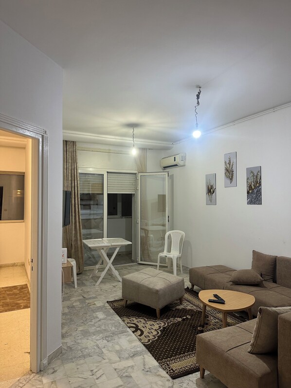 Una sala de estar moderna con un sofá, un reposapiés y una mesita. Hay una puerta de vidrio que da a un balcón con una mesa y sillas.
