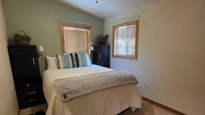 Un dormitorio con cama, ventilador de techo, mesita de noche, cómoda y ventana con persianas.
