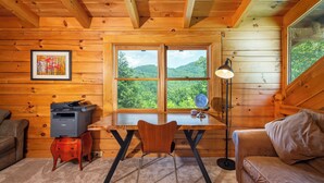 Interior de una cabaña de madera con escritorio, silla y sofá, un cuadro en la pared y una ventana que ofrece vistas a colinas verdes.