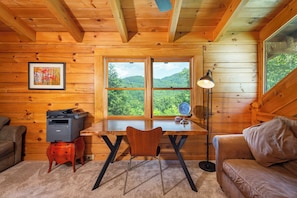 Ein Holzblockhaus-Interieur mit Schreibtisch, Stuhl und einer Couch, einem gerahmten Bild an der Wand und einem Fenster mit Blick auf grüne Hügel.