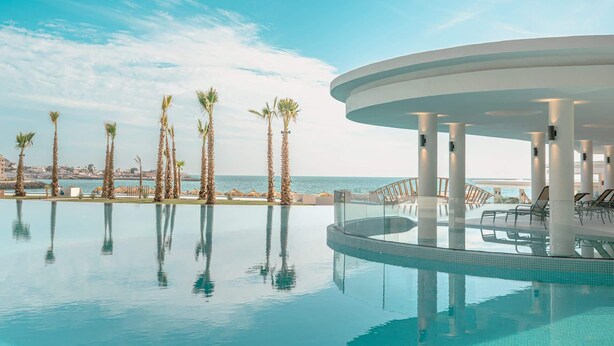 Un espace piscine agrémenté de palmiers, un salon fermé par des baies vitrées et une vue sur la mer.