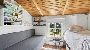 Ein Zimmer mit Holzboden, einem Bett mit gesteppter Bettdecke, einem grauen Sofa, einem Fenster mit Blick auf Grünflächen und Regalen mit Büchern und Dekoration.