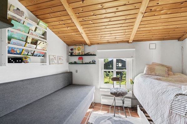 Ein Zimmer mit Holzboden, einem Bett mit gesteppter Bettdecke, einem grauen Sofa, einem Fenster mit Blick auf Grünflächen und Regalen mit Büchern und Dekoration.