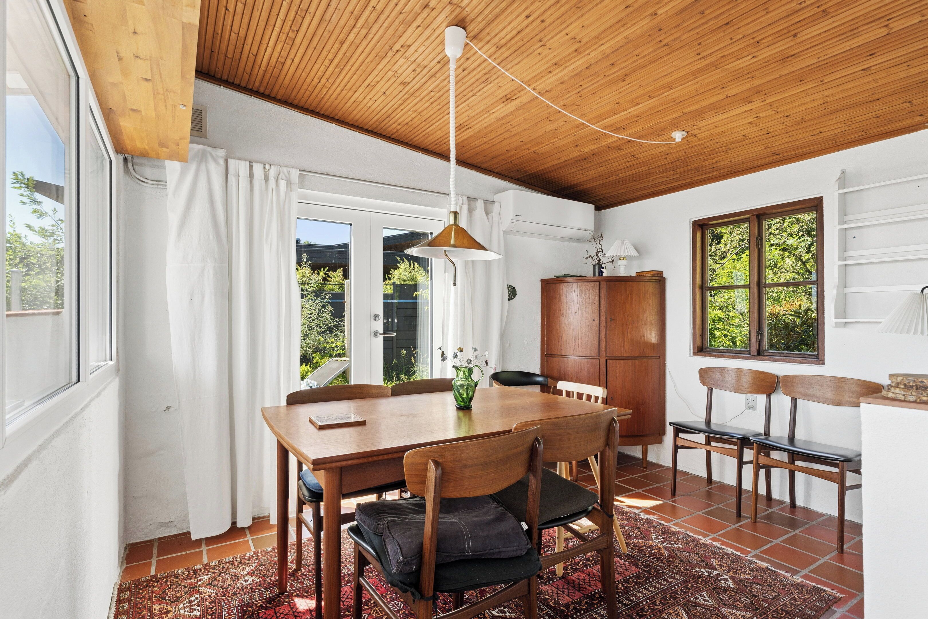 Comedor con mesa de madera, sillas, lámpara colgante y vistas al exterior con vegetación.