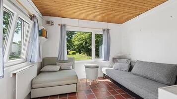 Une pièce avec un plafond en bois, une fenêtre avec des rideaux, un radiateur, une petite table, un canapé gris et un fauteuil vert.