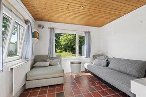 A room with a wooden ceiling, a window with curtains, a radiator, a small table, a grey sofa, and a green armchair.