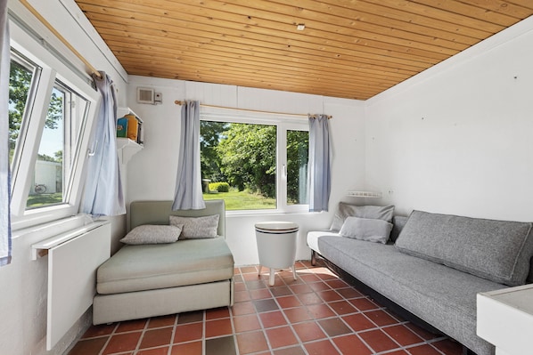 A room with a wooden ceiling, a window with curtains, a radiator, a small table, a grey sofa, and a green armchair.
