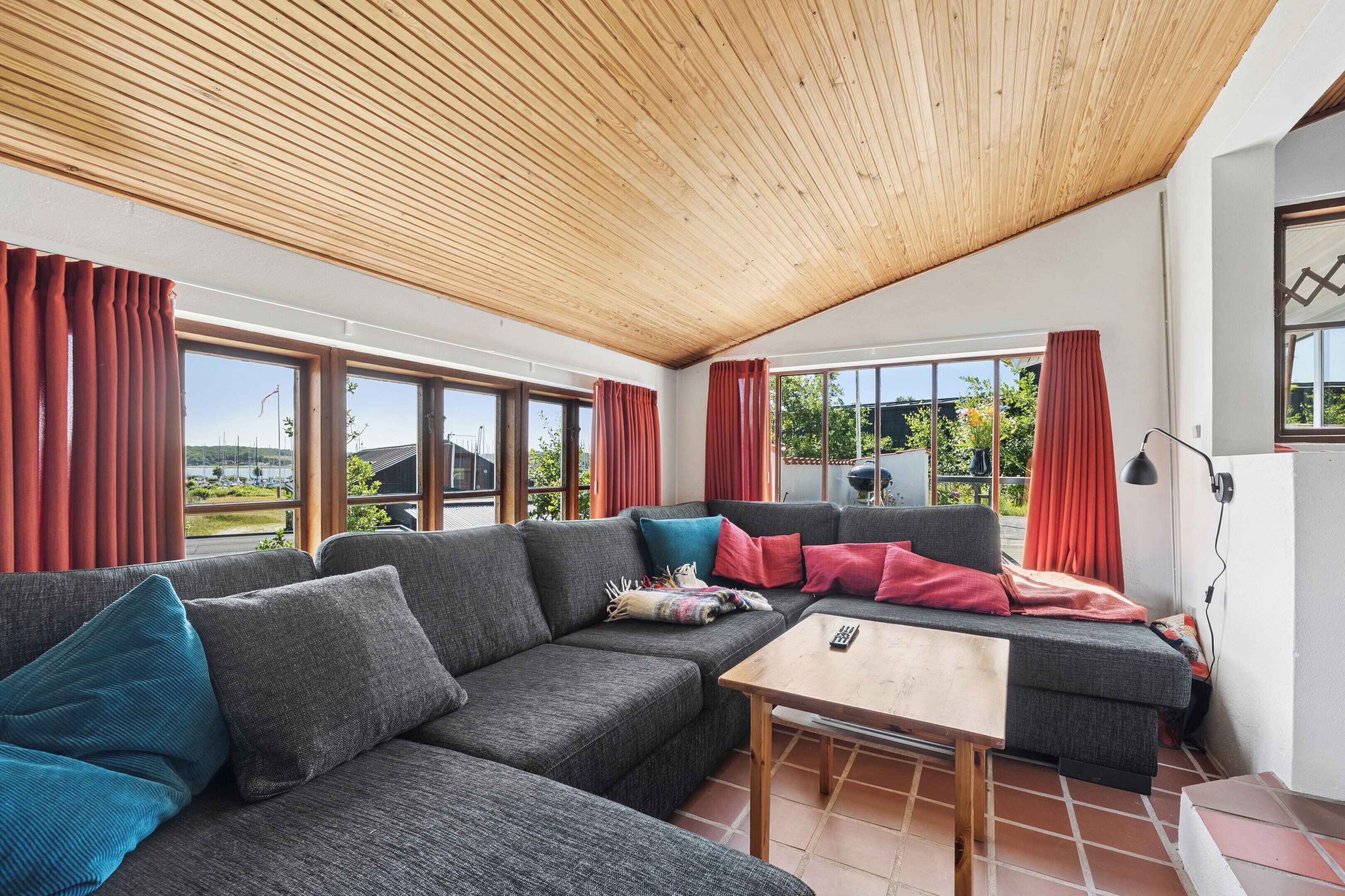 A modern living room with a wooden ceiling, large windows, and a sofa with multiple cushions.